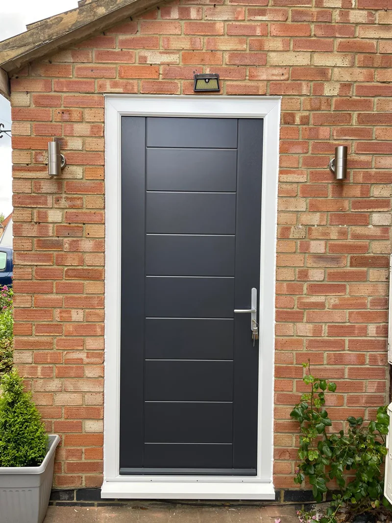An anthracite grey composite front door with three horizontal grooved panels and a full-height brushed handle, in a plain red-brick porch.