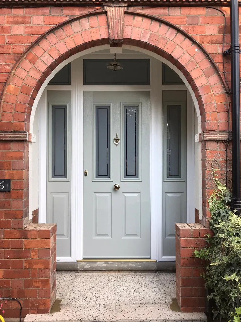 A light grey composite front door with a glazed upper panel, fitted into the arched brick opening of a porch with planting to one side.