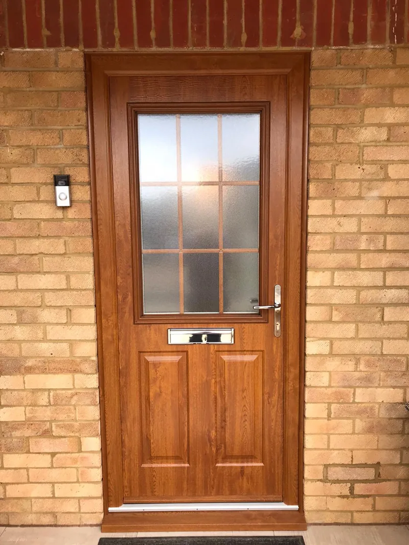An oak-effect woodgrain composite front door with a square glazed panel and a wall lantern beside it, in a red-brick elevation.