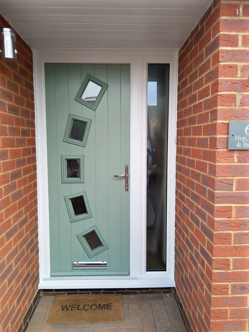 A sage green composite front door with two tall glazed panels and a matching side panel, fitted in a red-brick porch with a coir mat on the step.